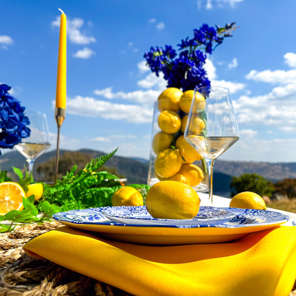 close up image of lemon placed on plate with yellow napkin, yellow candle and blue flowers